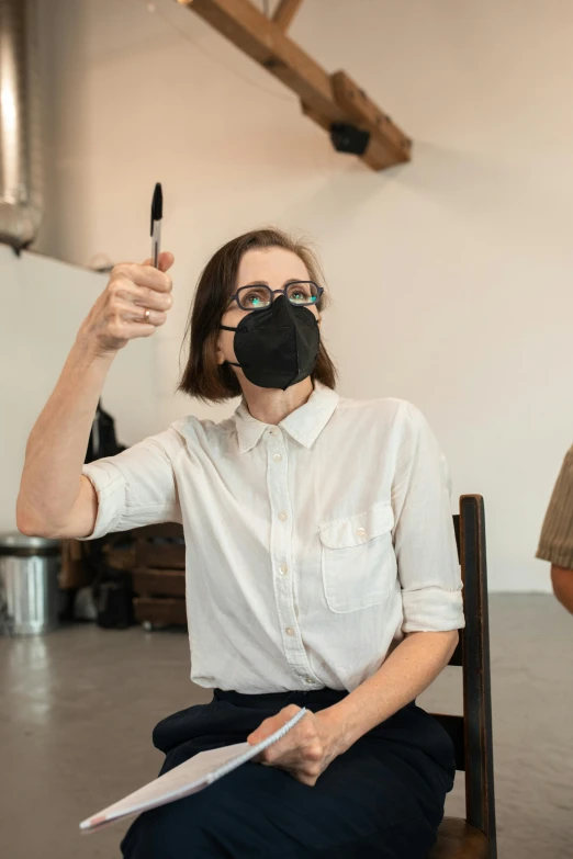 a woman sitting on a chair wearing a face mask, arbeitsrat für kunst, holding a small vape, wearing black rimmed glasses, 2019 trending photo, in a workshop