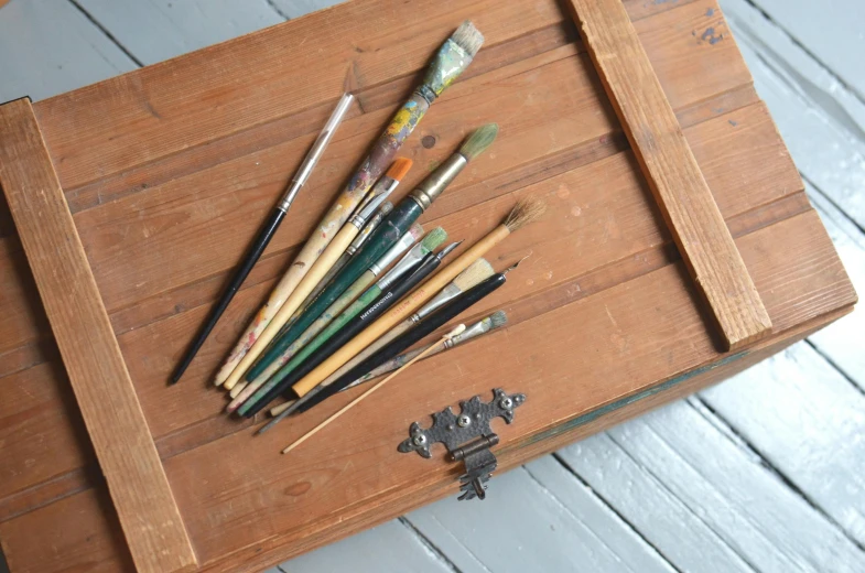 a bunch of paint brushes sitting on top of a wooden box, a detailed painting, by Sylvia Wishart, unsplash, academic art, vintage 1960 print, easel, brown, military storage crate