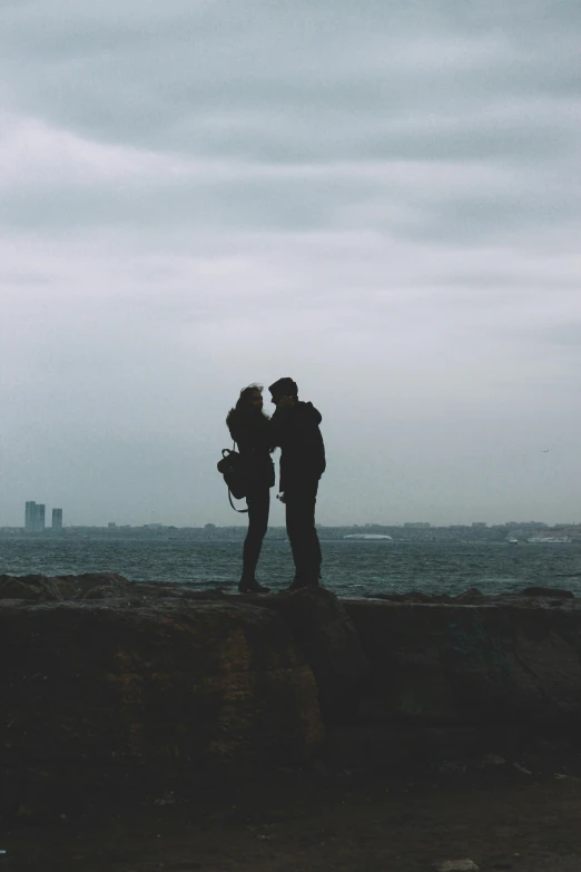 a couple standing on top of a cliff next to the ocean, gloomy skies, silhouetted, surrounding the city, making out