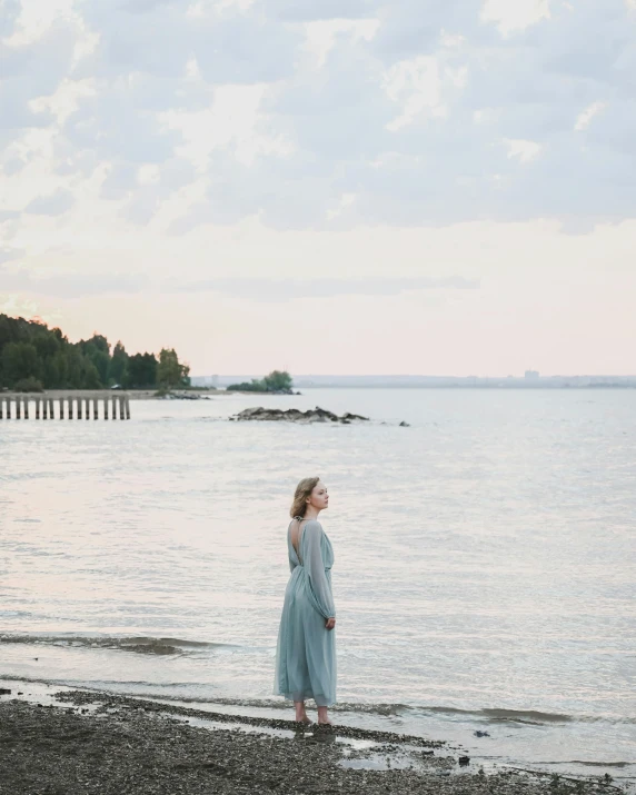 a woman standing on a beach next to a body of water, vancouver, in muted colors, midsommar style, in the evening