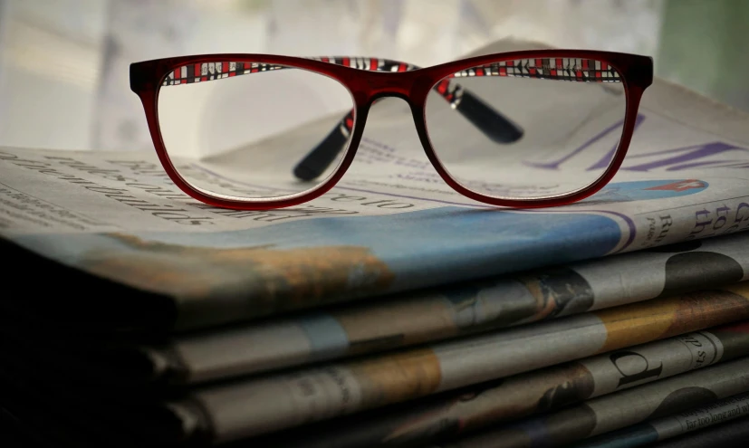 a pair of glasses sitting on top of a stack of newspapers, a picture, wearing red tainted glasses, intricate details in the frames, press shot, medium close shot