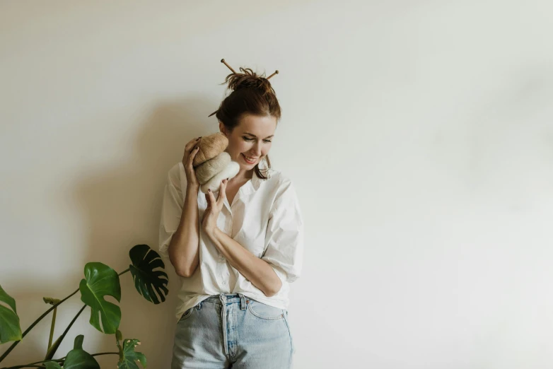 a woman standing next to a potted plant, by Emma Andijewska, minimalism, brown hair in two buns, smiling playfully, with a wooden stuff, promo image