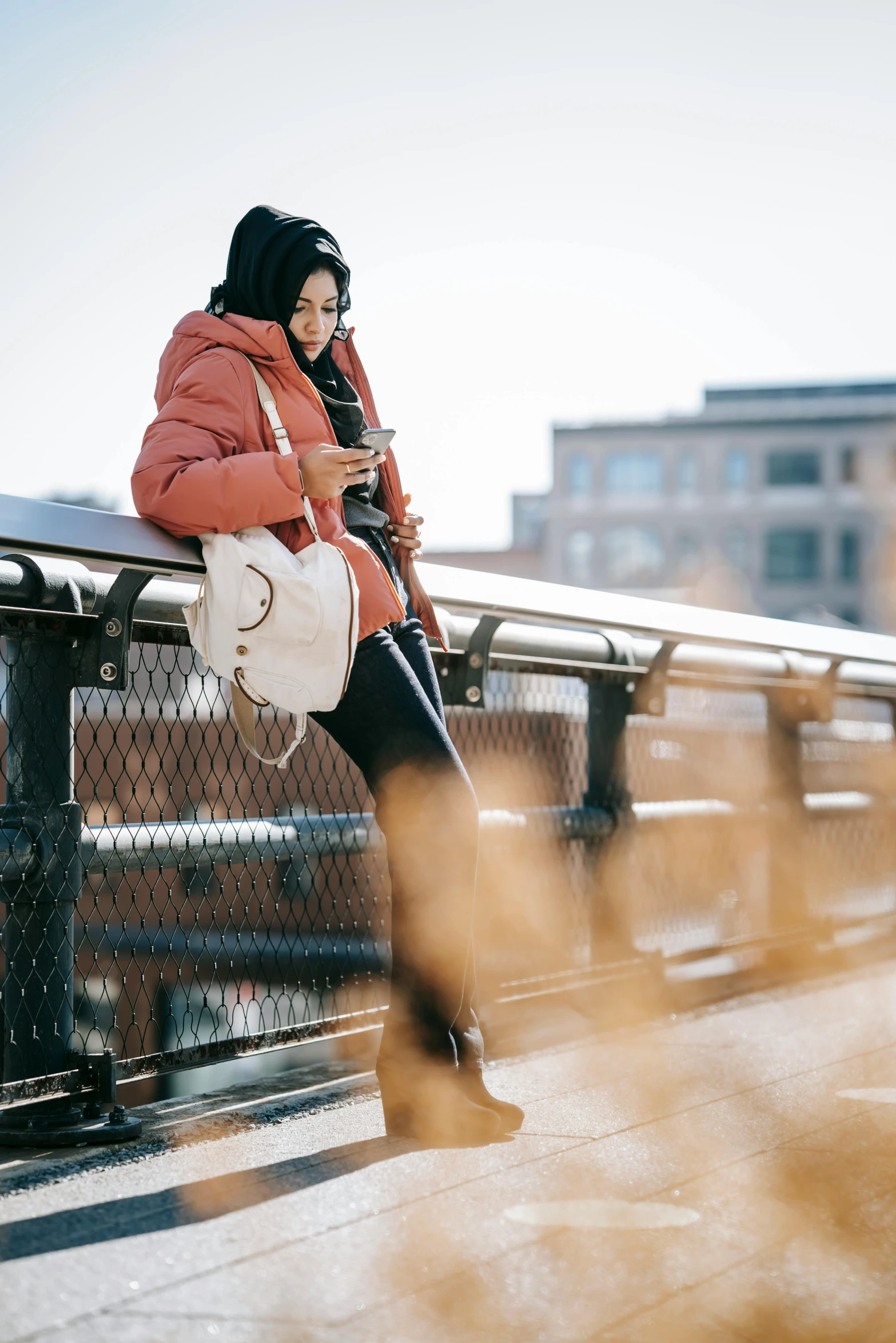 a woman sitting on a fence looking at her cell phone, trending on pexels, happening, orange reflective puffy coat, hijab, walking, swedish