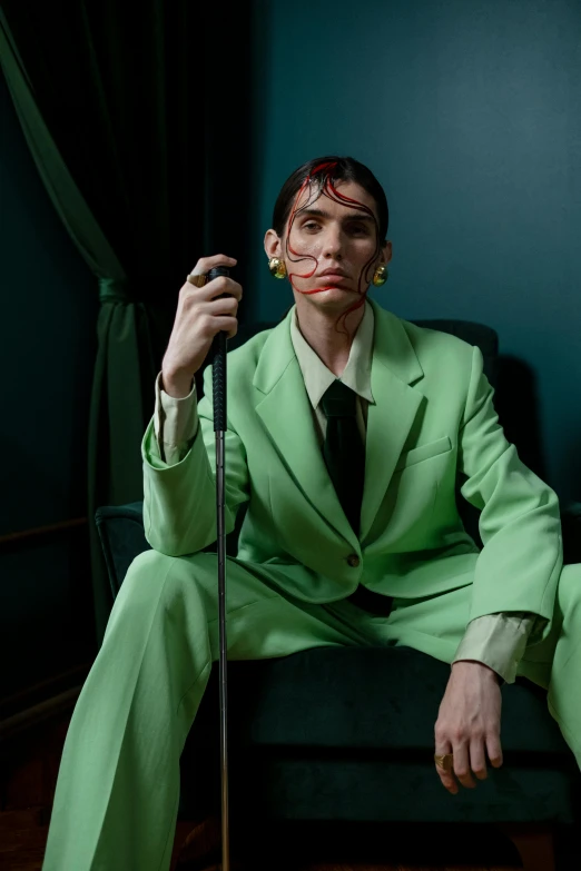 a man in a green suit sitting on a chair, by Caro Niederer, trending on pexels, bauhaus, androgynous vampire, non binary model, joker makeup, thom browne