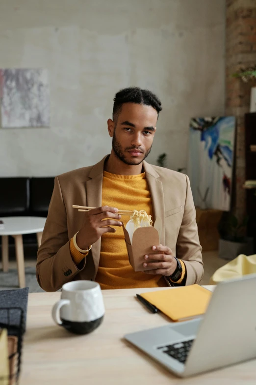 a man sitting at a table with a laptop and a cup of coffee, eating noodles, in a gold suit, gen z, tailored clothing