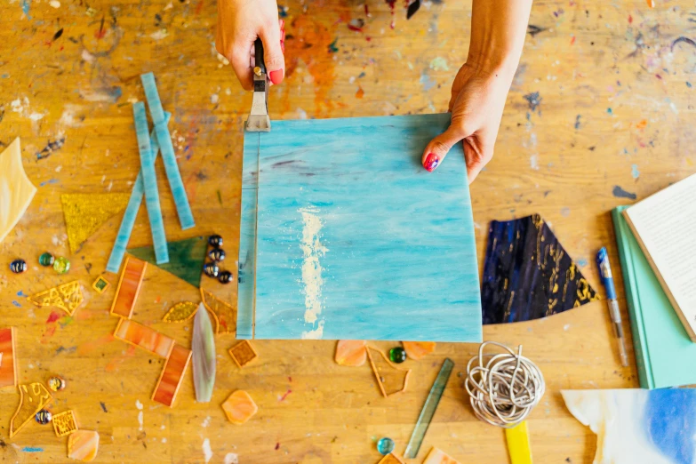 a person holding a piece of paper on top of a table, a photorealistic painting, by Jessie Algie, trending on unsplash, process art, cyan and gold scheme, colorful glass art, paint knife, a wooden