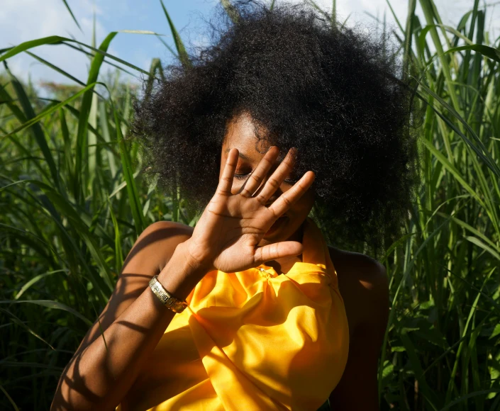 a woman covering her face with her hands, by Lily Delissa Joseph, pexels contest winner, afrofuturism, long thick grass, wavy hair yellow theme, afro, summer light