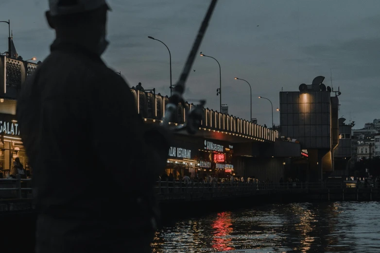 a man standing next to a body of water holding a fishing pole, by Attila Meszlenyi, pexels contest winner, watching night streets, advertising photo, seen from the side, bridge