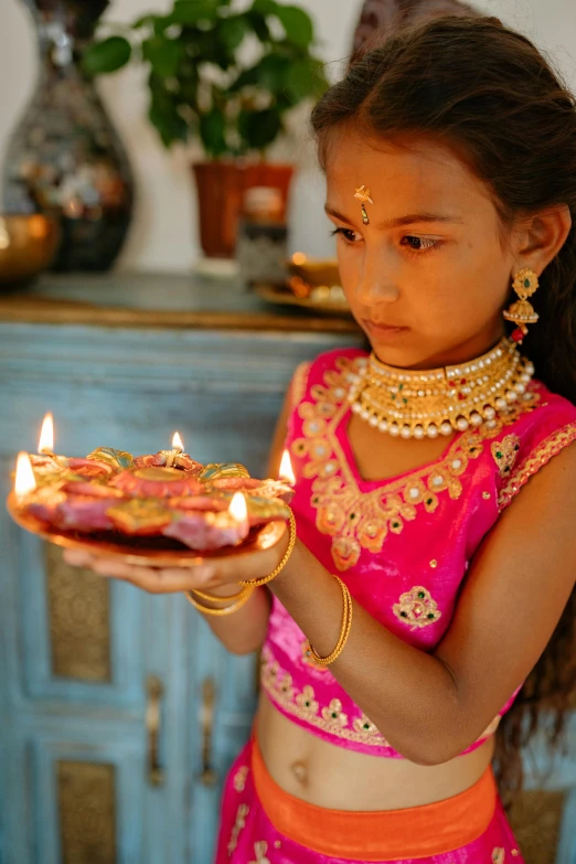 a little girl holding a cake with lit candles, by Alice Mason, hurufiyya, wearing gold jewellery, indian style, summer lighting, jeweled ornament over forehead