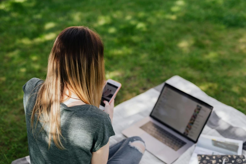 a woman sitting on a blanket with a laptop and cell phone, trending on pexels, sitting in the garden, computer screens, student, back - shot