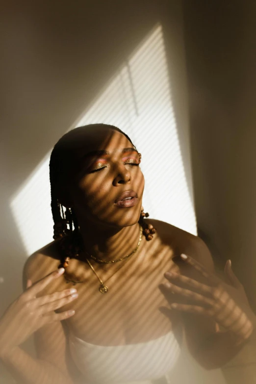 a woman in a white dress standing in front of a window, an album cover, by Lily Delissa Joseph, trending on pexels, light and space, praying at the sun, with brown skin, underexposed lighting, she has a glow coming from her