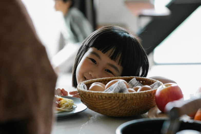 a little girl sitting at a table with a bowl of food, inspired by Yukimasa Ida, pexels contest winner, eggs, fruits in a basket, profile image, asian face