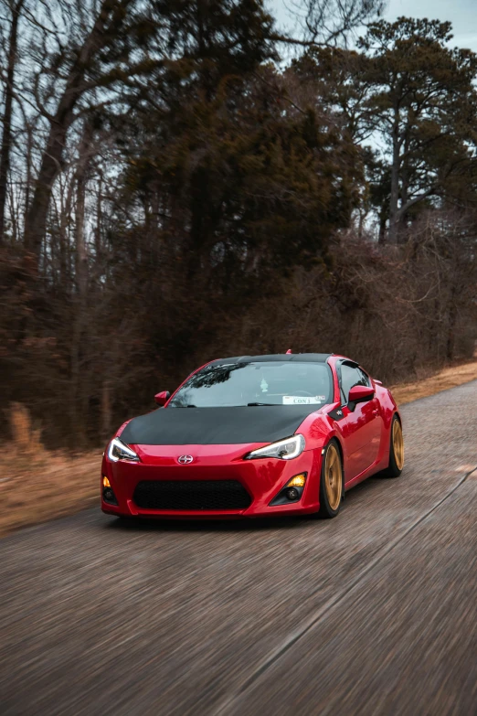 a red car driving down a road next to a forest, a picture, by Jason Felix, unsplash, samurai vinyl wrap, full - body - front - shot, golden detailing, soft top