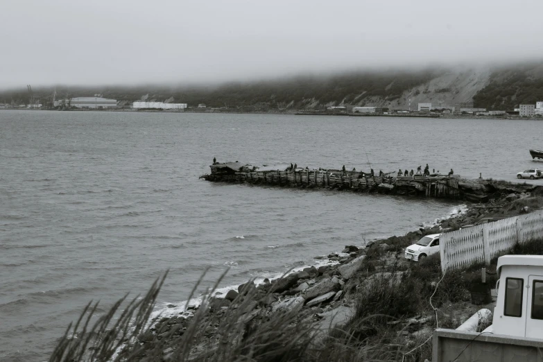 a boat sitting on top of a body of water, a black and white photo, under a gray foggy sky, wood pier and houses, inuit heritage, mermaids in distance