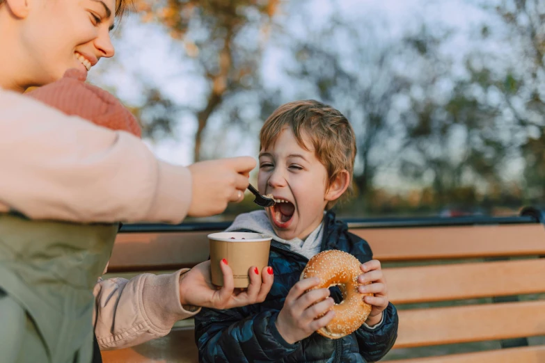 a woman feeding a child a donut while sitting on a bench, pexels contest winner, manuka, with a cup of hot chocolate, lachlan bailey, owen klatte