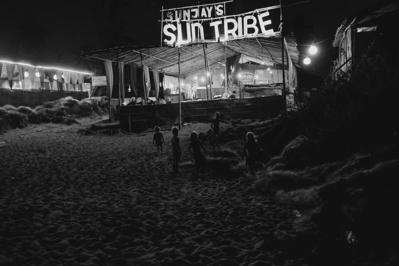 a group of people standing on top of a sandy beach, an album cover, sunshafts, tribals, it's night time, black and white picture