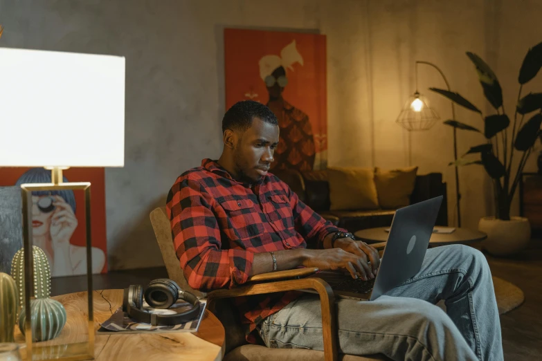 a man sitting in a chair using a laptop, a portrait, trending on pexels, ( ( dark skin ) ), red shirt brown pants, sitting across the room, early evening