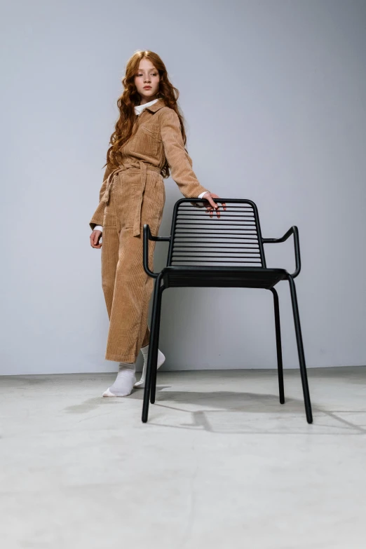 a woman standing next to a chair in a room, by Nina Hamnett, pexels contest winner, altermodern, wearing human air force jumpsuit, roots and hay coat, matte black pants, made of wire
