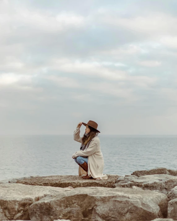 a woman sitting on top of a rock next to the ocean, straw hat and overcoat, lgbtq, 2019 trending photo, wearing a cardigan