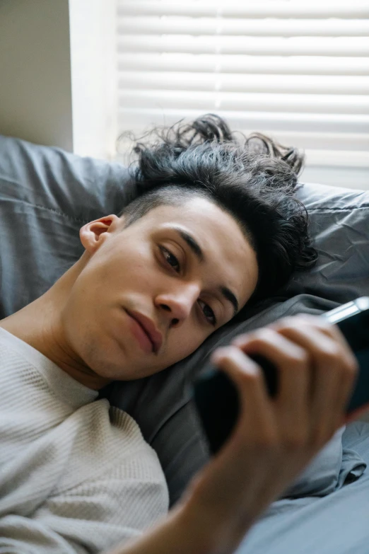 a man laying in bed using a cell phone, trending on pexels, happening, boy has short black hair, non binary model, concerned, long spiky hair