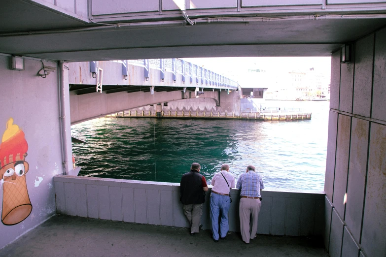 a group of people standing next to a body of water, a picture, under bridge, looking around a corner, espoo, manly