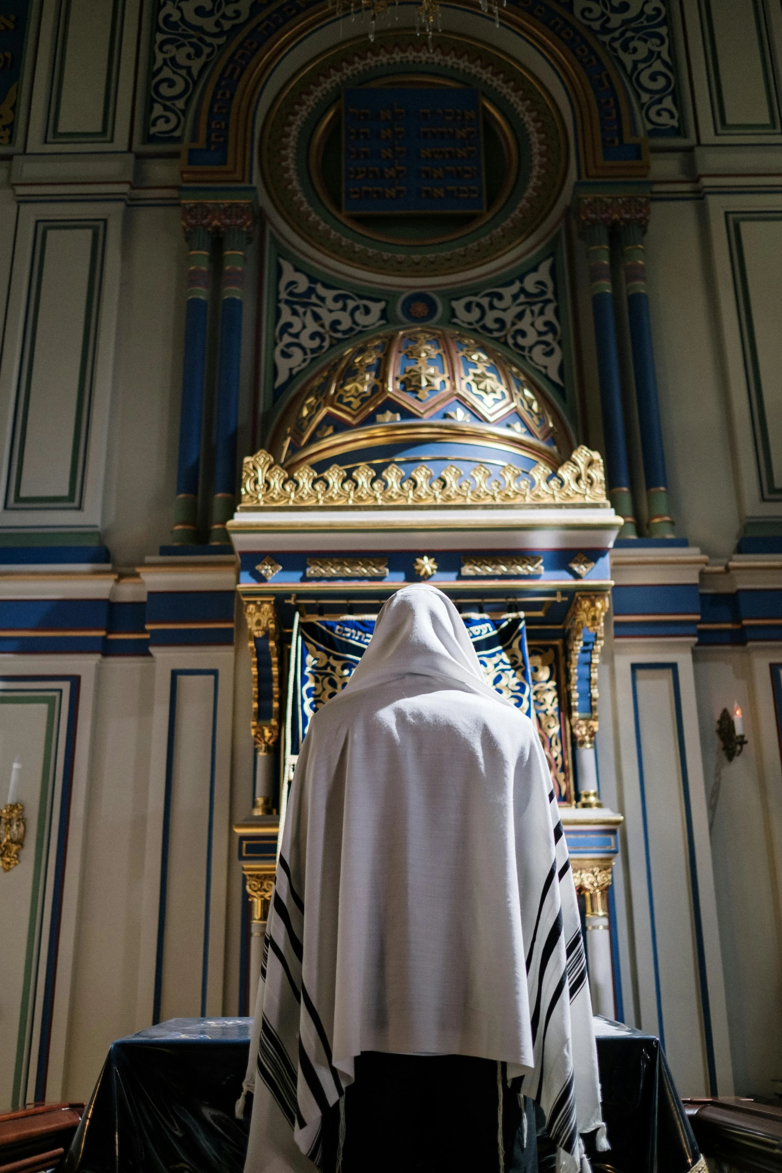 a priest standing at the alter of a church, by Julia Pishtar, symbolism, mosque synagogue interior, wearing white silk hood, white + blue + gold + black, facing away from camera