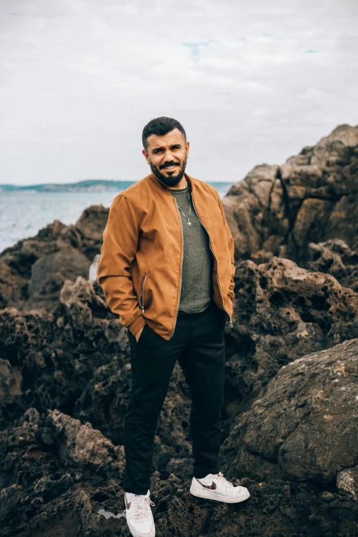 a man standing on rocks near the ocean, an album cover, inspired by Oluf Høst, pexels contest winner, wearing brown leather jacket, he got a big french musctache, a portrait of rahul kohli, sydney hanson