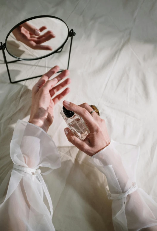 a woman holding a bottle of perfume in front of a mirror, inspired by Elsa Bleda, trending on pexels, visual art, white bed, lit from above, wearing a white blouse, hands with five fingers