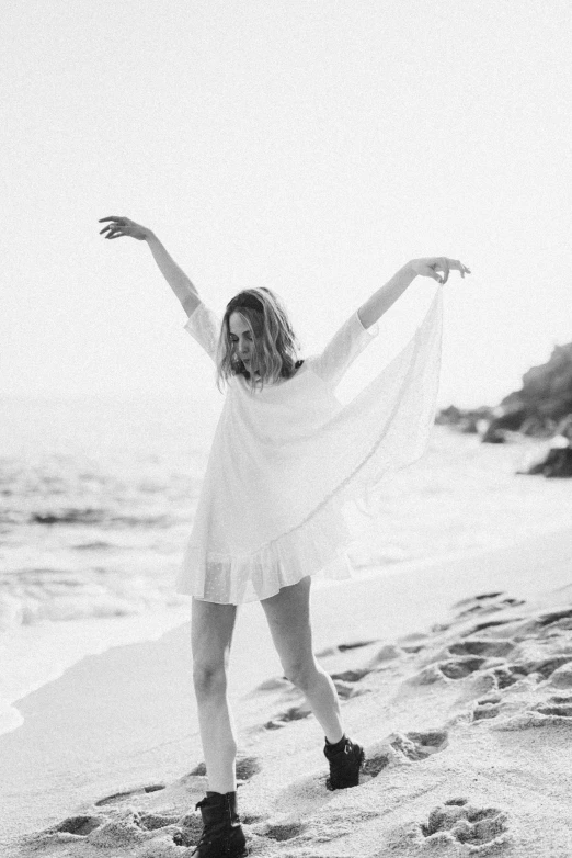 a woman standing on top of a beach next to the ocean, a black and white photo, happening, girl in white dress dancing, photoshoot for skincare brand, wearing torn white cape, wave a hand at the camera