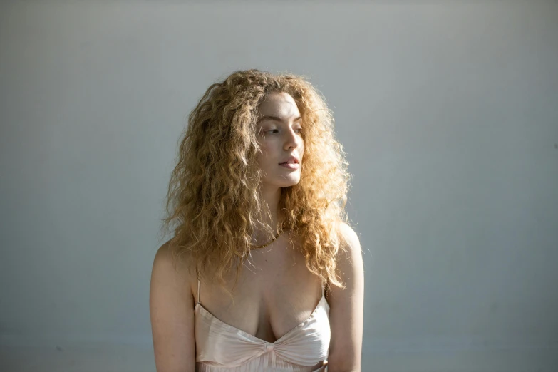 a beautiful young woman sitting on top of a bed, an album cover, unsplash, photorealism, pale skin curly blond hair, diffuse natural sun lights, standing in a dimly lit room, chest up bust shot