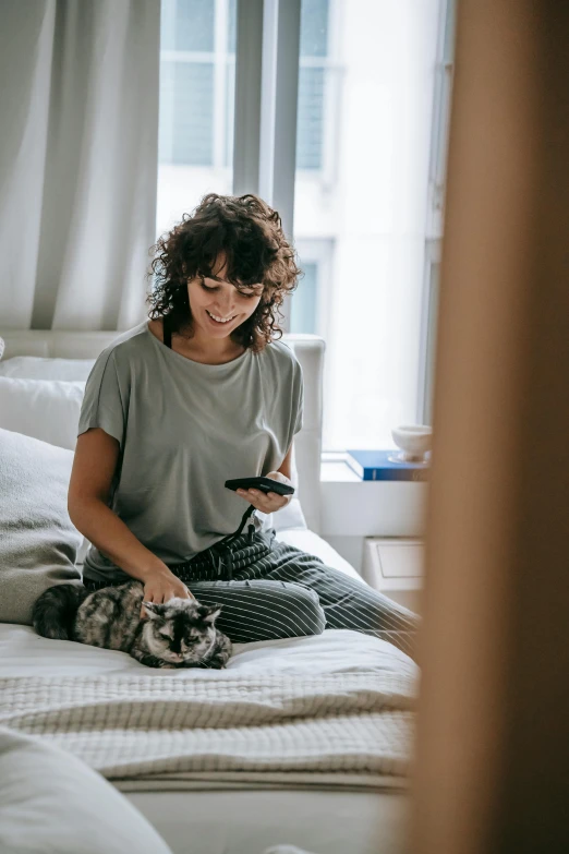 a woman sitting on a bed looking at her phone, a cat is smiling, watching tv, shot with sony alpha, cat on top of dog