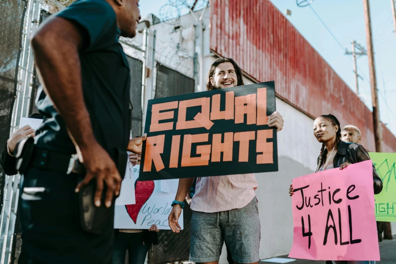 a group of people holding signs in front of a building, by Julia Pishtar, trending on pexels, feminist art, gay rights, background image, standing in a township street, a person standing in front of a