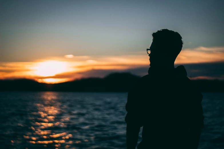 a person standing in front of a body of water, silhouette of a man, man with glasses, sunset in the distance, profile picture 1024px