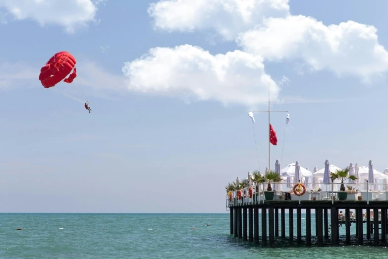 a group of people standing on top of a pier flying kites, by Niyazi Selimoglu, red flags holiday, as an air balloon, seaview, on stilts