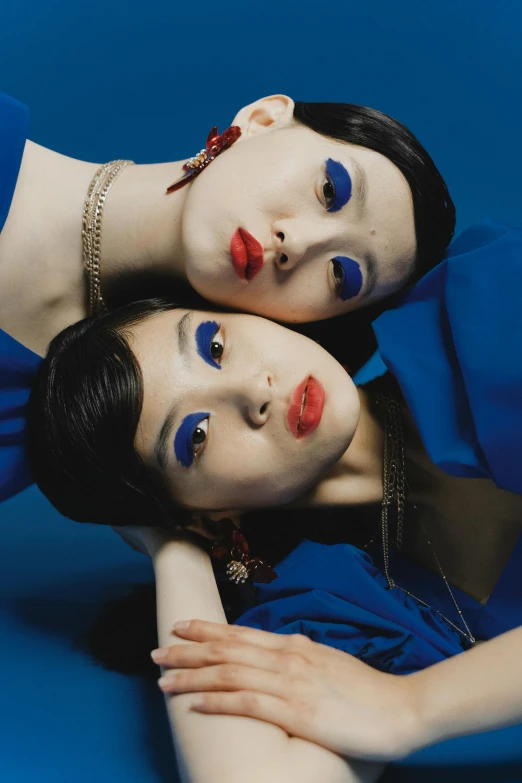 two women laying next to each other on a blue surface, an album cover, inspired by Fei Danxu, dada, kabuki makeup, dark blue lipstick, cobalt blue and pyrrol red, asian face