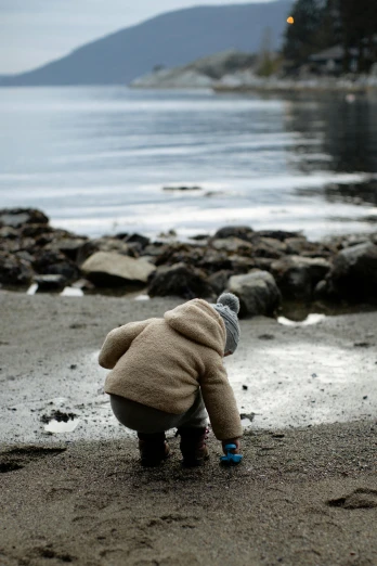 a person kneeling on a beach near a body of water, a picture, toddler, head down, humans exploring, wandering