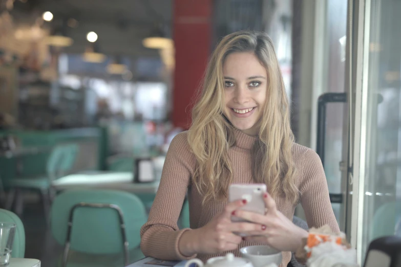 a woman sitting at a table looking at her cell phone, pexels contest winner, young blonde woman, youtube thumbnail, cute smile, in classic diner