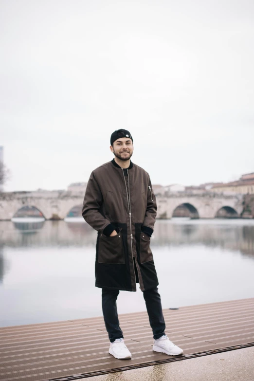 a man standing in front of a body of water, zayn malik, brown tuffle coat, in paris, portait image