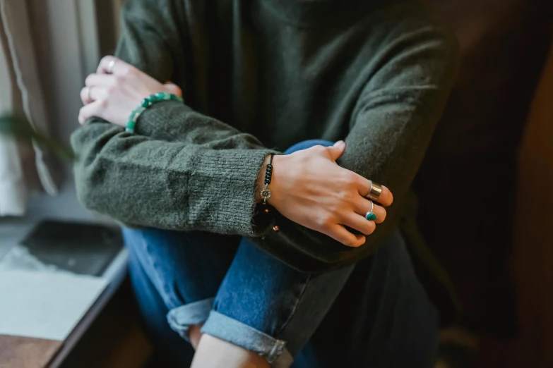 a woman sitting on top of a couch next to a window, by Emma Andijewska, trending on pexels, bracelets, wearing a green sweater, holding each other hands, androgynous male