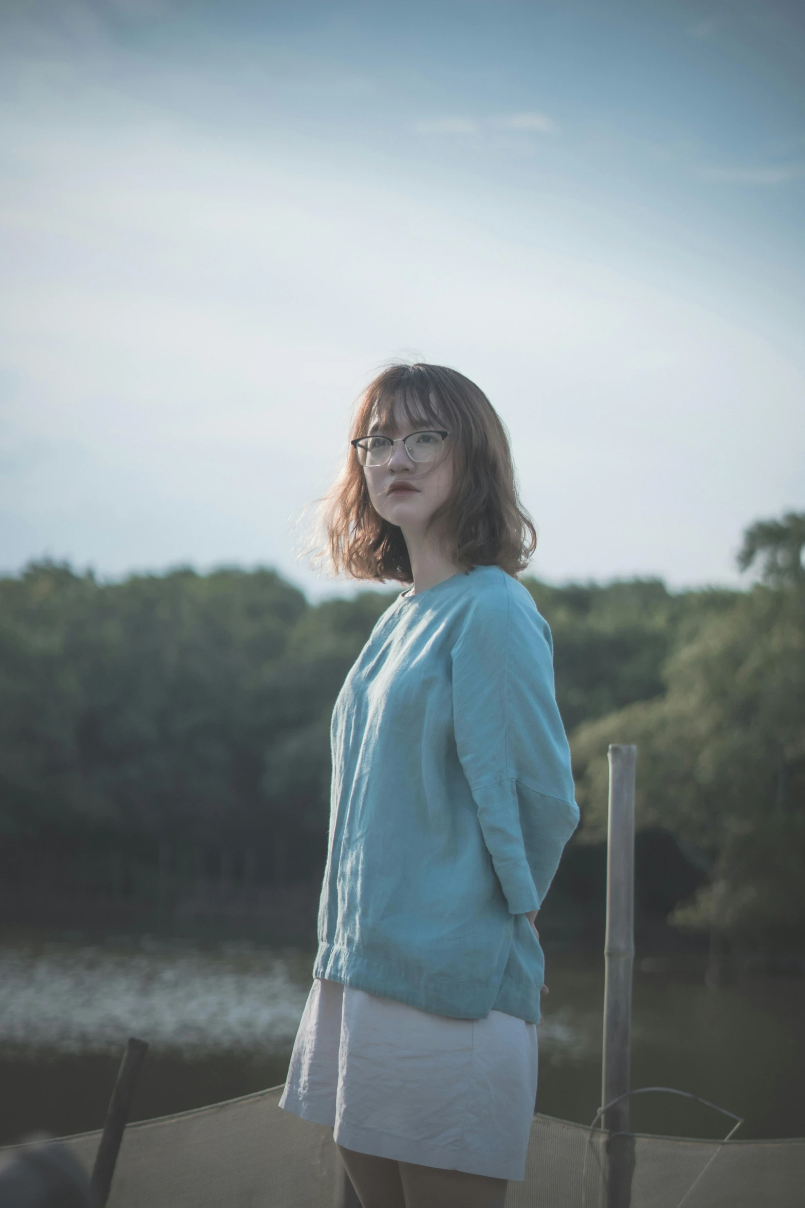 a woman standing on a dock next to a body of water, by Attila Meszlenyi, blue sweater, girl wearing round glasses, ((portrait)), low light cinematic