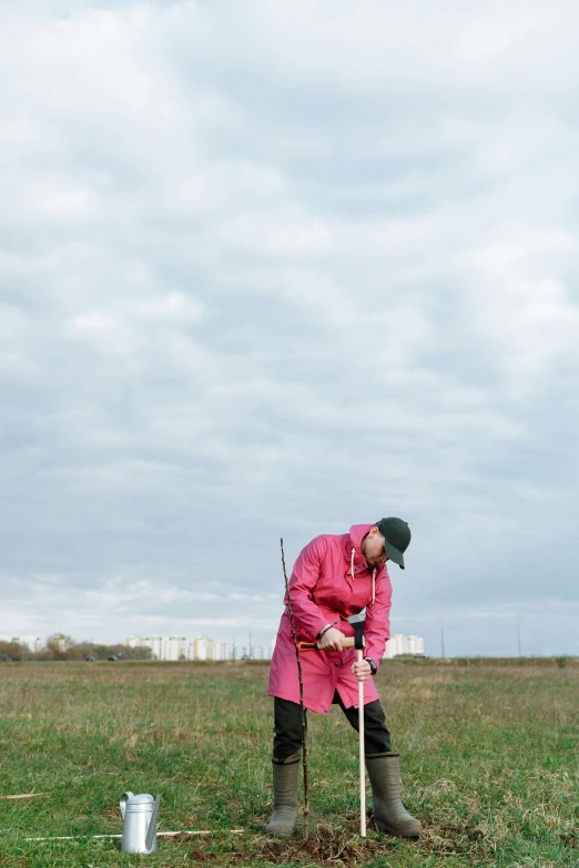 a woman standing in a field with a shovel, by Attila Meszlenyi, unsplash, happening, wearing a pink hoodie, in russia, golf course, grazing
