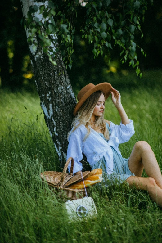 a woman sitting in the grass next to a tree, by Adam Marczyński, pexels contest winner, straw hat, picnic, instagram model, early evening