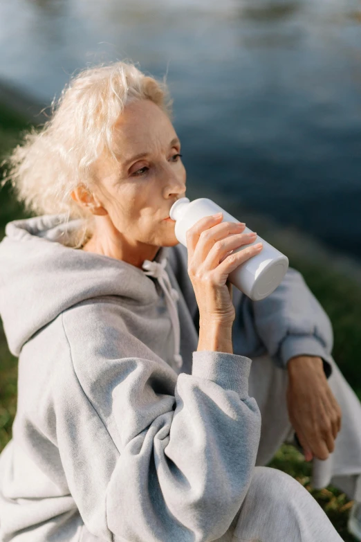 a woman sitting next to a body of water, holding a bottle, white haired lady, hydration, profile image