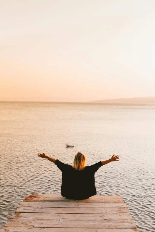 a woman sitting on a dock with her arms outstretched, trending on unsplash, minimalism, red sea, celebration, praying, overlooking the beach