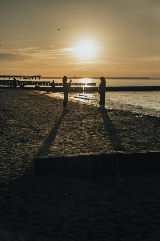 a couple of people standing on top of a sandy beach, tallinn, casting long shadows, the sun is setting, brown