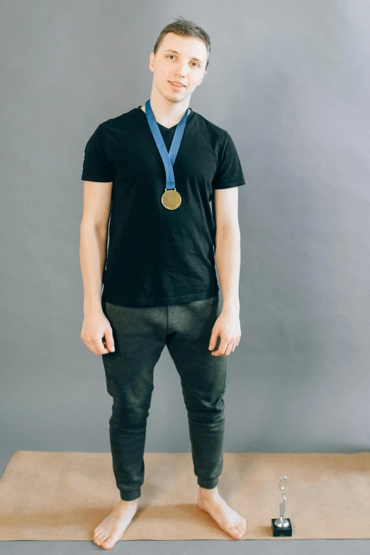a man standing on a mat with a medal around his neck, inspired by Alexander Kanoldt, pexels contest winner, wearing pants and a t-shirt, plain background, skinny caucasian young man, a person standing in front of a