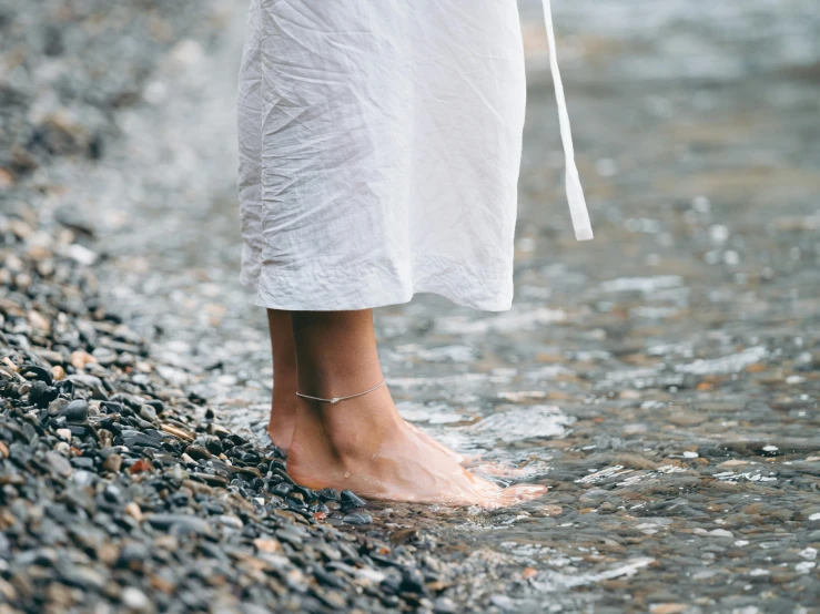 a person standing on a beach next to a body of water, trending on unsplash, foot wraps, sparkling in the flowing creek, wearing a white hospital gown, sleek legs