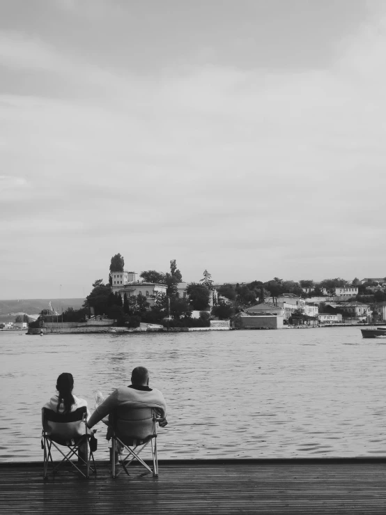 a couple of people sitting next to a body of water, a black and white photo, by Zofia Stryjenska, small castle in the distance, rostov, 15081959 21121991 01012000 4k