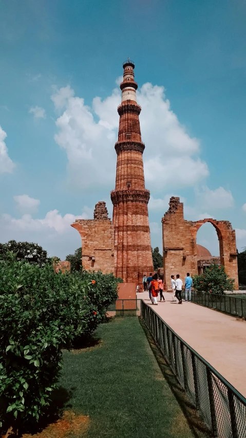 a tall tower sitting on top of a lush green field, by Bernardino Mei, pexels contest winner, renaissance, ancient india, brick building, people walking around, examining ruins