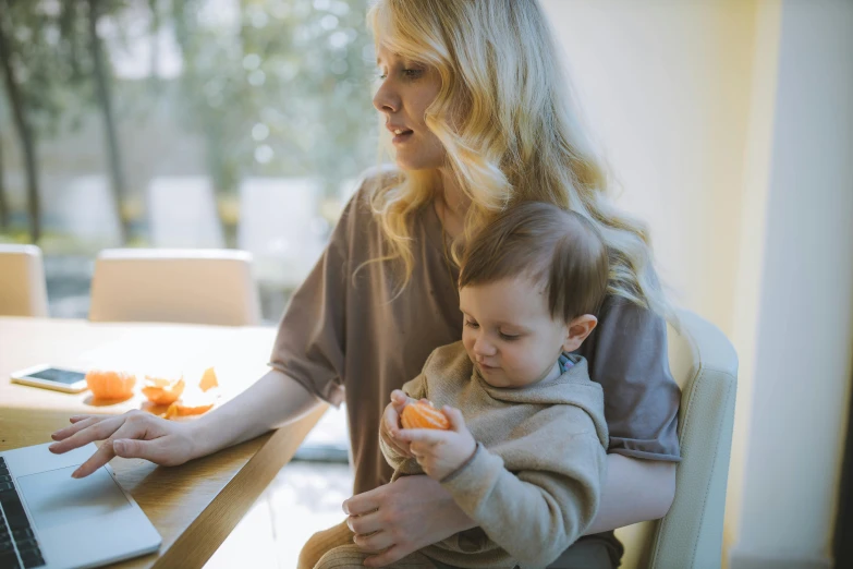 a woman holding a baby in front of a laptop, by Julian Hatton, pexels, holding a tangerine, blonde, avatar image, sitting at the table